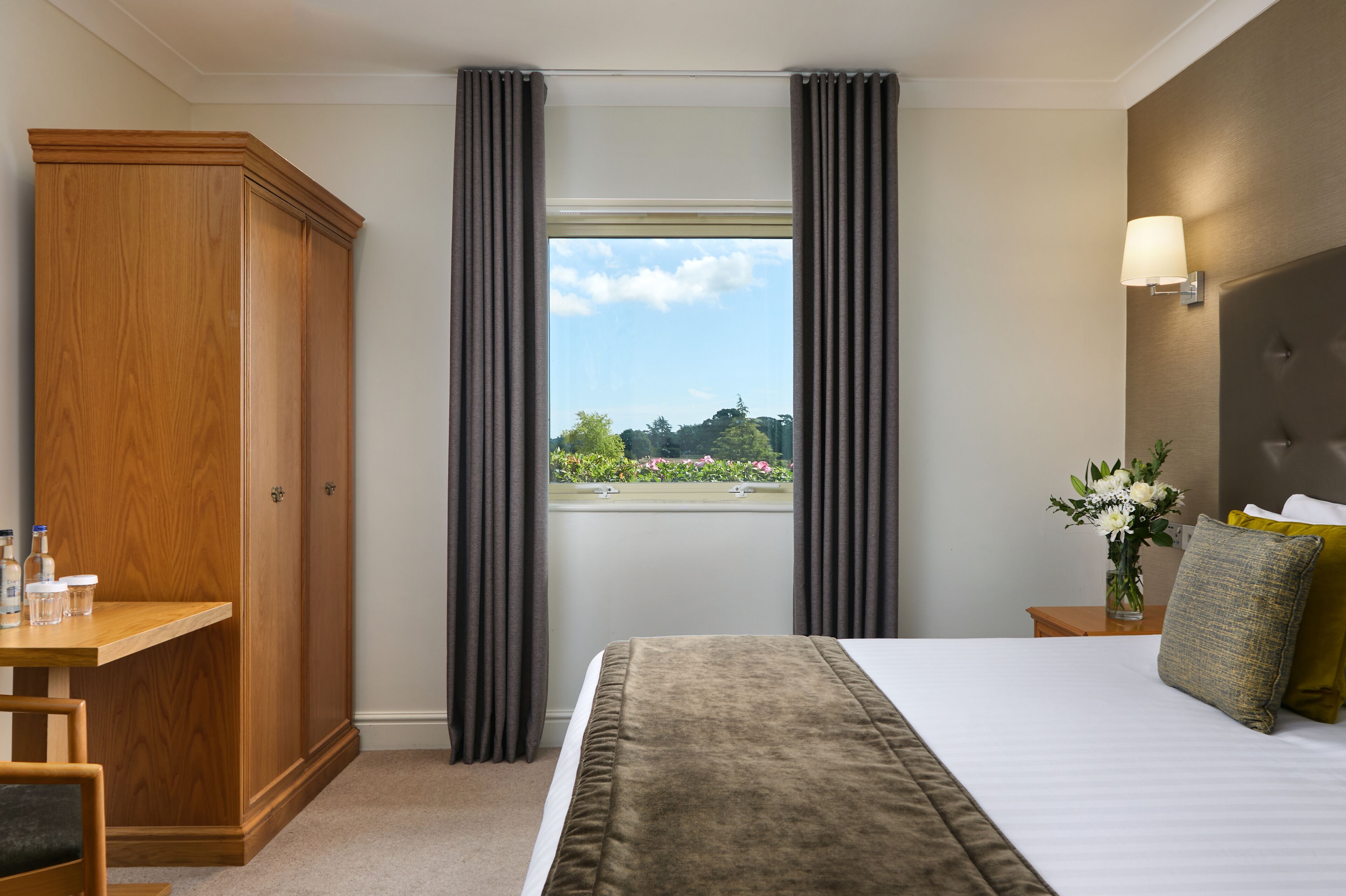 A hotel room with a large bed, a wardrobe, a bedside table with a vase of flowers, and a window with curtains.