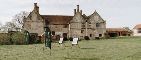 A large, historic stone building with multiple chimneys and windows, surrounded by a grassy field with two target boards set up.