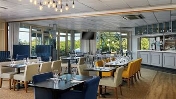 A restaurant with multiple tables and chairs, a bar area, and large windows offering a view of the outdoors.