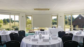 A conference room with tables set for a meeting, large windows offering a view of the outdoors, and a projector screen at the front.