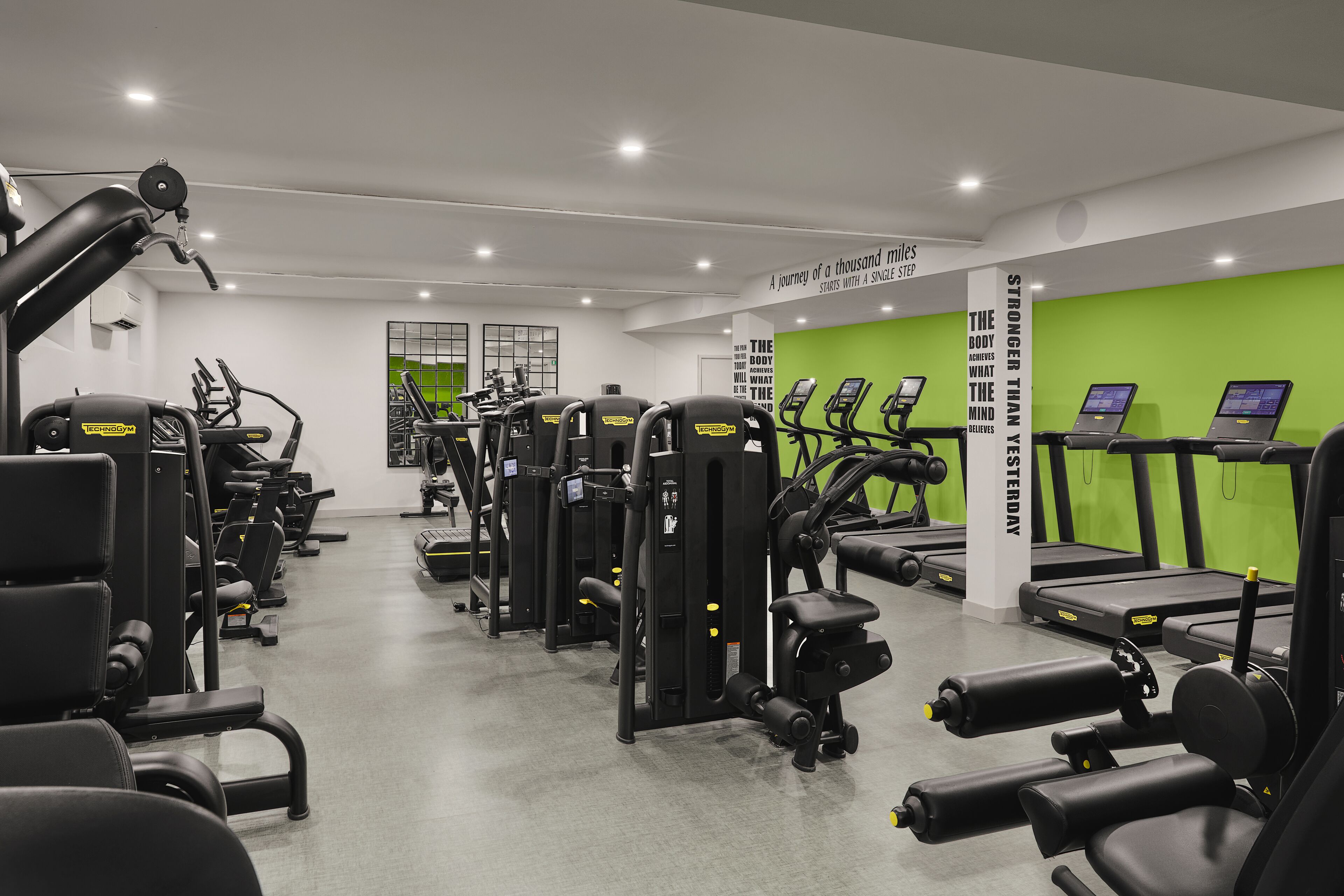 A modern gym with various exercise machines, treadmills, and a green wall with motivational quotes.