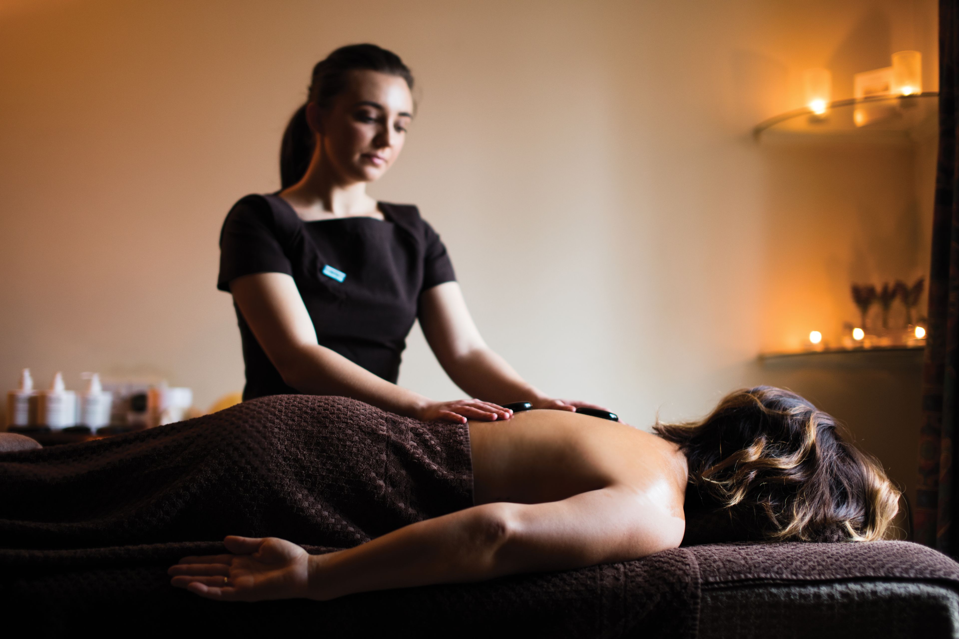 En massagebehandling i et rum med en seng, massagestol og en vægmonteret hylde med flasker.