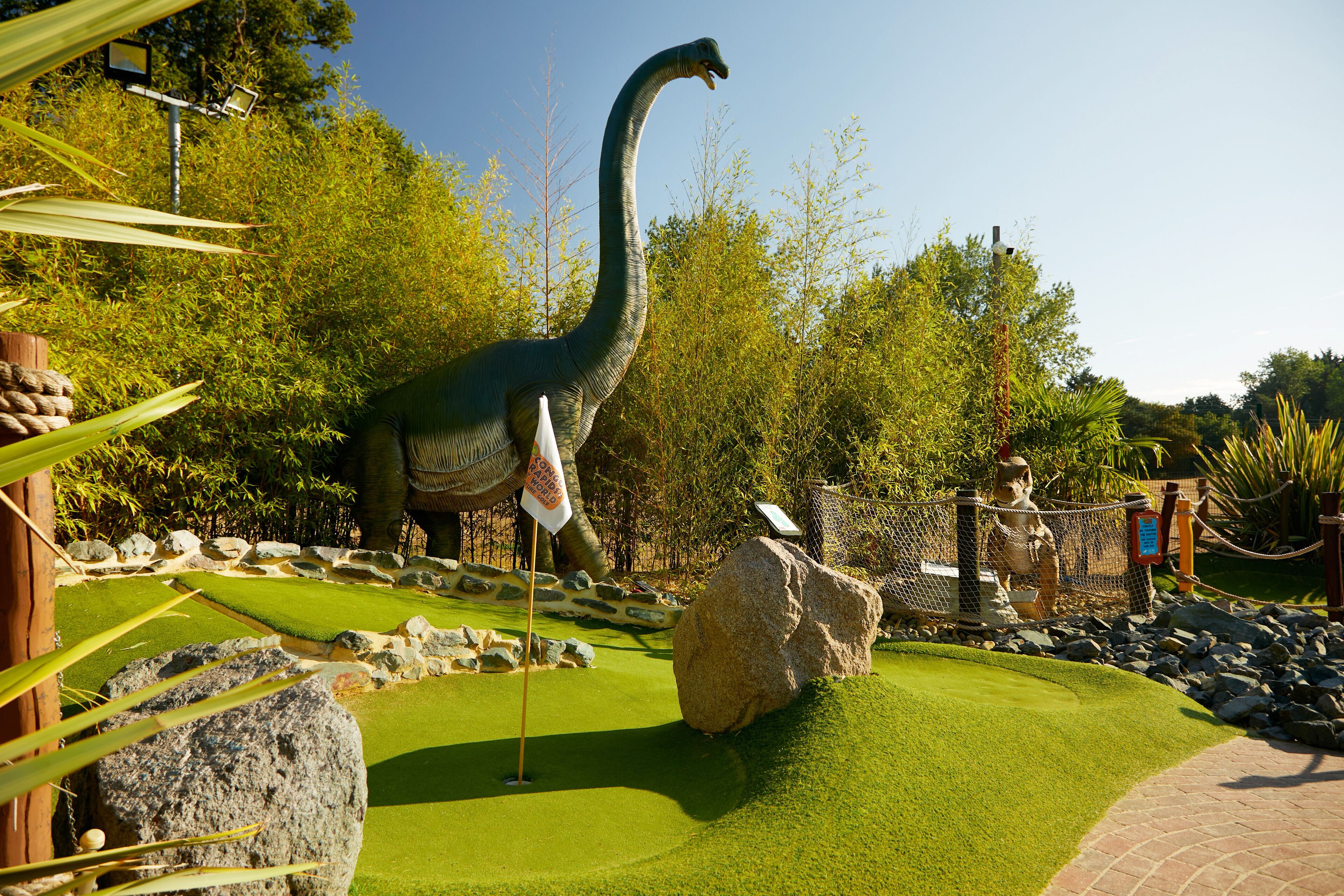 Une statue de dinosaure dans un espace aménagé avec des rochers, un drapeau et une clôture.