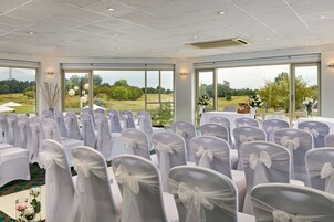 Un salón de conferencias con filas de sillas blancas decoradas con lazos, amplias ventanas que dan a un campo de golf y una mesa con un arreglo floral.