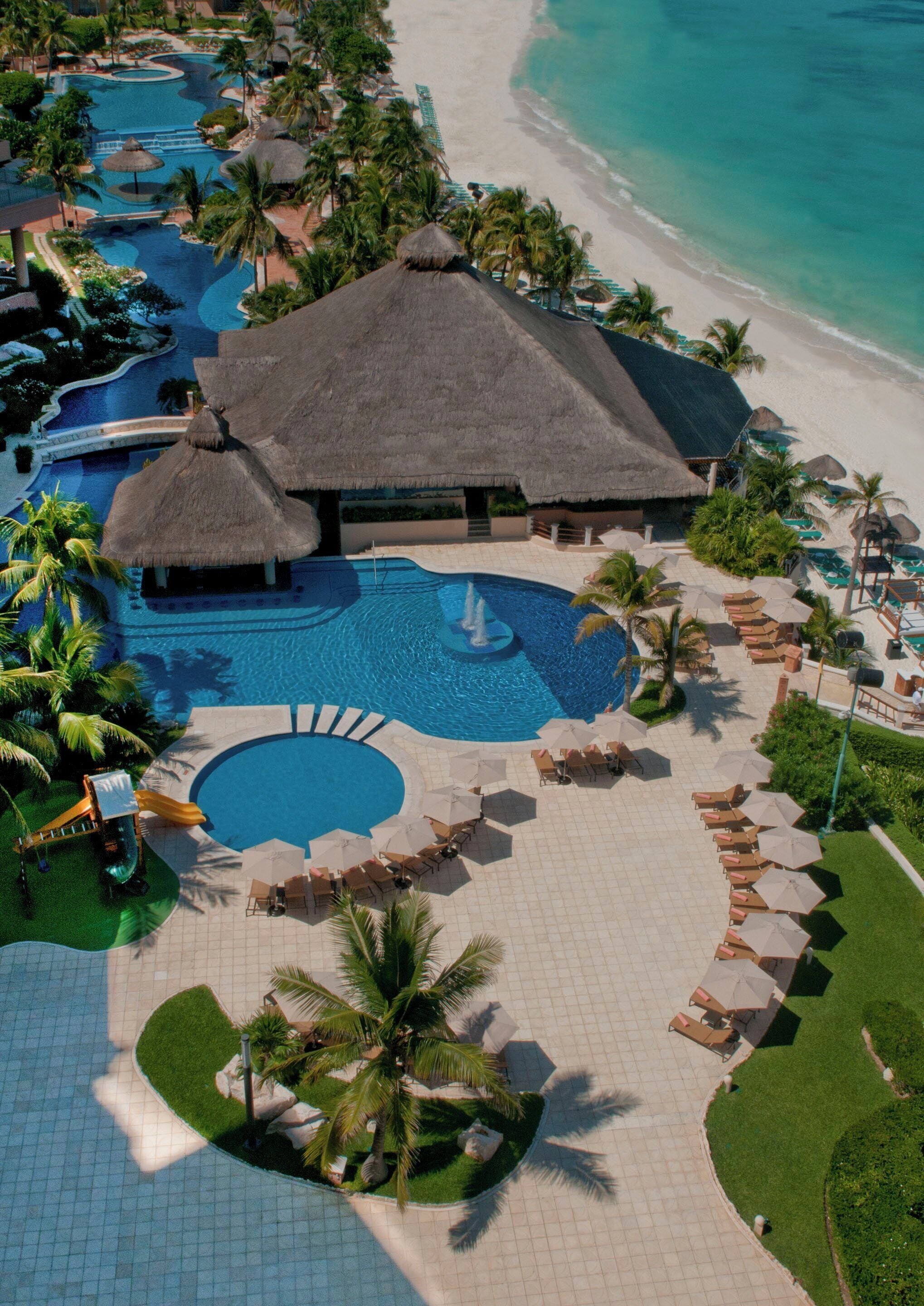 An aerial view of a resort with a large pool area, palm trees, and a beach.