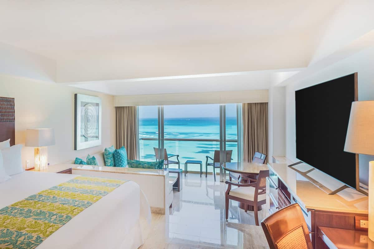 A hotel room with a large bed, a desk with a chair, a TV, and a view of the ocean.