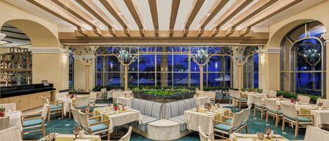 Un vaste espace de restauration intérieur doté d’un coin salon central, de plusieurs tables et chaises, ainsi que de grandes fenêtres agrémentées d’éléments décoratifs.
