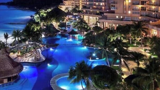 A resort with multiple swimming pools, a large building with balconies, and a beach in the background.