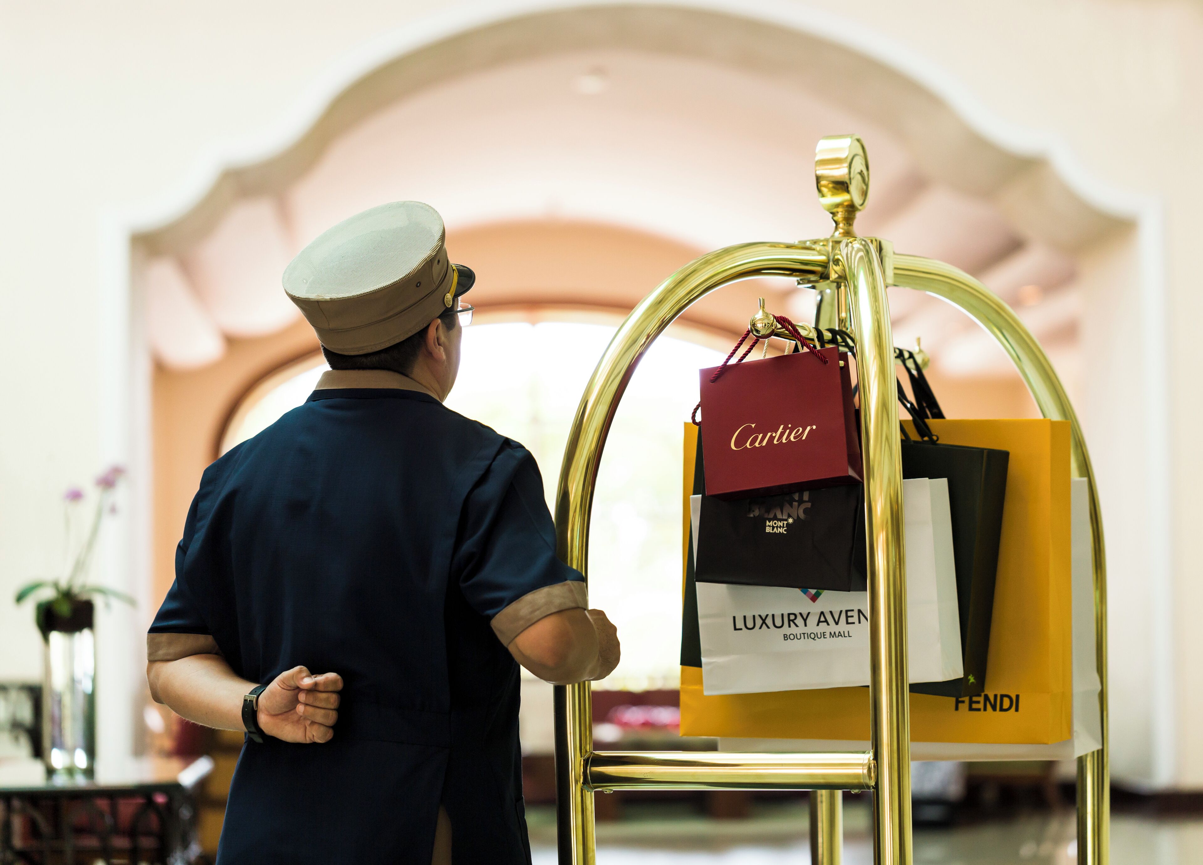 A man in a uniform standing next to a gold-colored shopping cart with luxury brand bags.