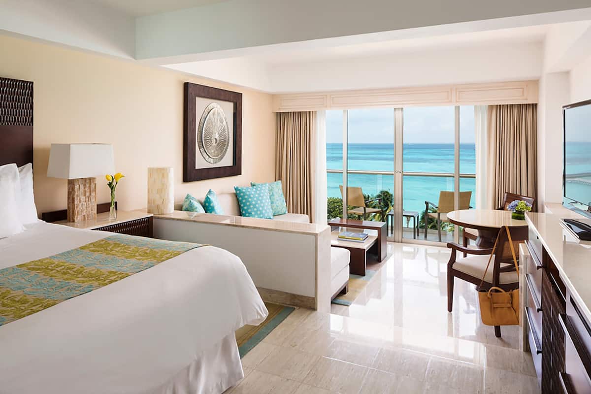 A hotel room with a bed, a desk, a chair, a balcony with a view of the ocean, and a television.