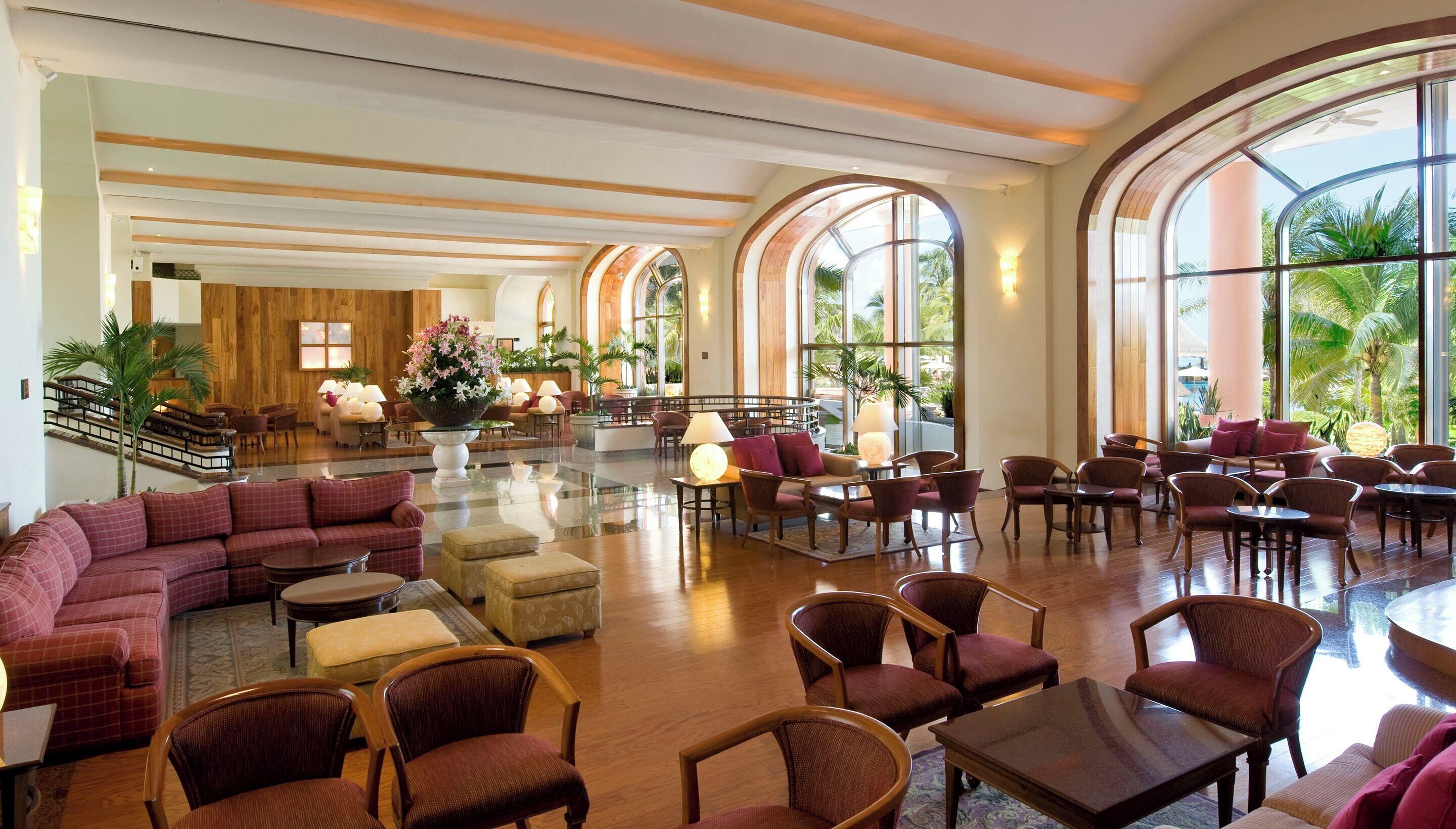 A spacious lobby with a central seating area, multiple tables, and large arched windows offering views of palm trees.