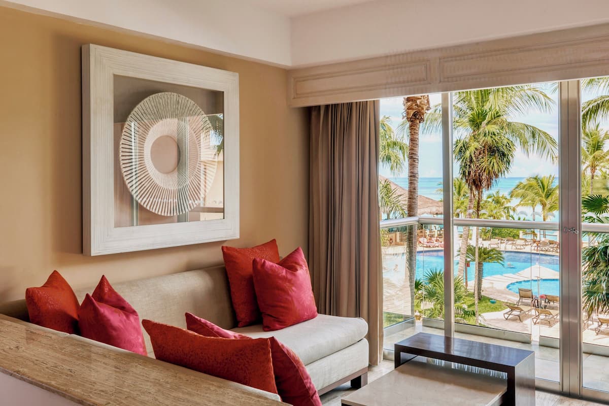 A room with a sofa, red cushions, a framed artwork, and a view of a pool and palm trees.