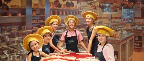 Un grupo de personas en una cocina con ollas de cobre colgando del techo y un mantel rojo sobre la mesa.