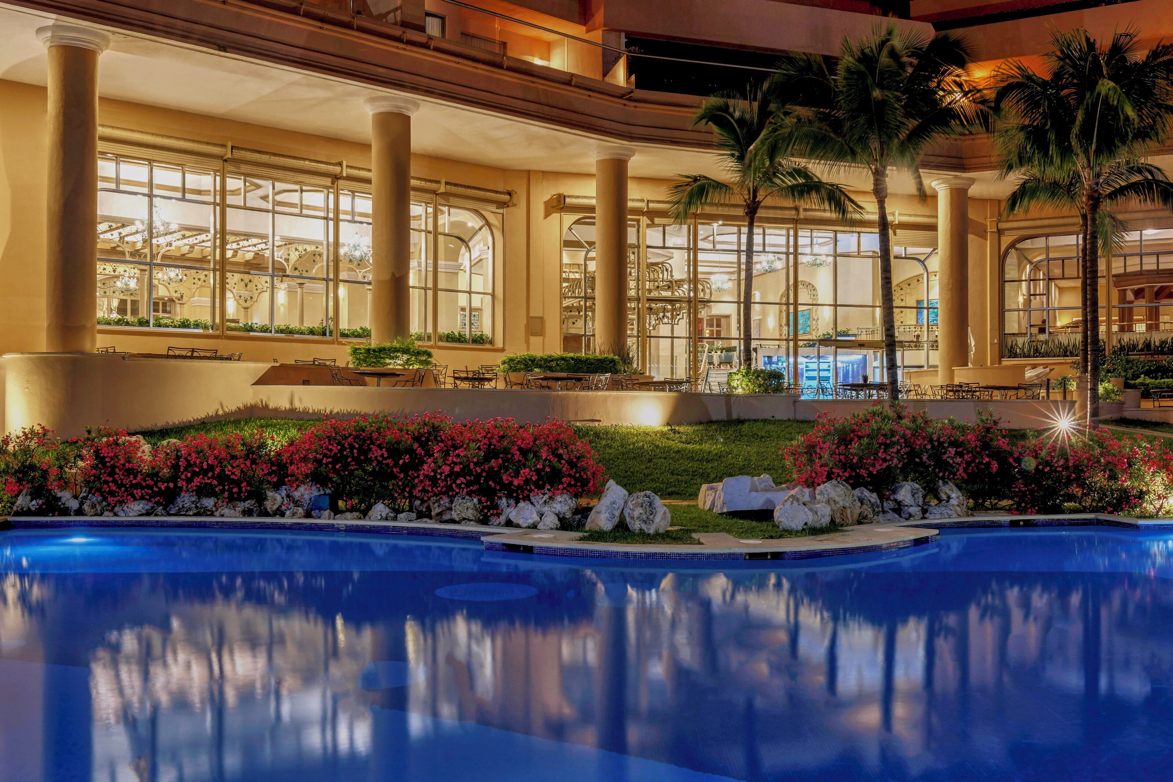 Zona de piscina con el reflejo de la arquitectura del edificio, rodeada de palmeras y plantas con flores.