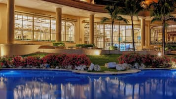 A pool area with a reflection of the building's architecture, surrounded by palm trees and flowering plants.