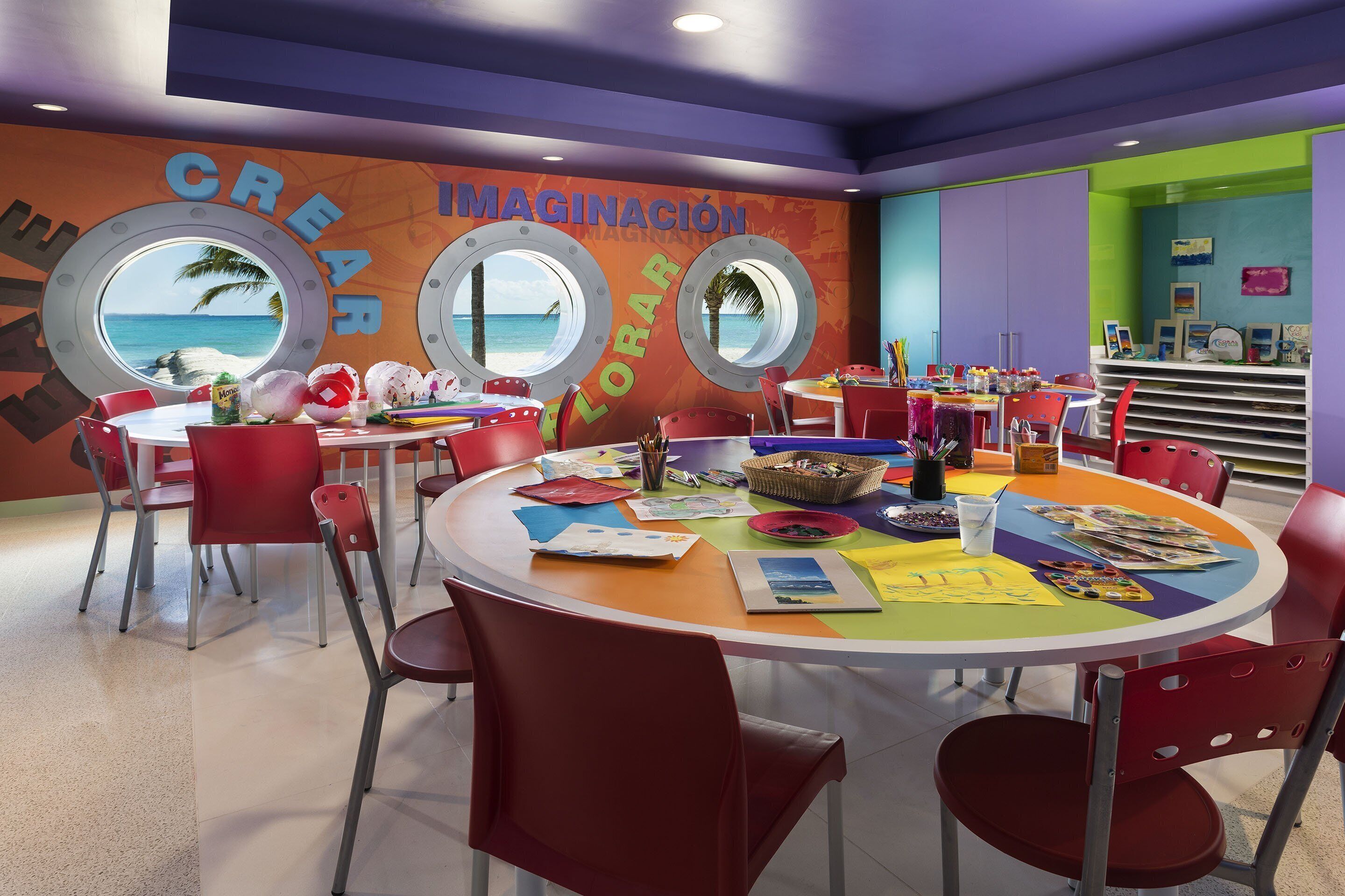 Una habitación colorida con ventanas redondas, sillas rojas y varios materiales de arte sobre una mesa.