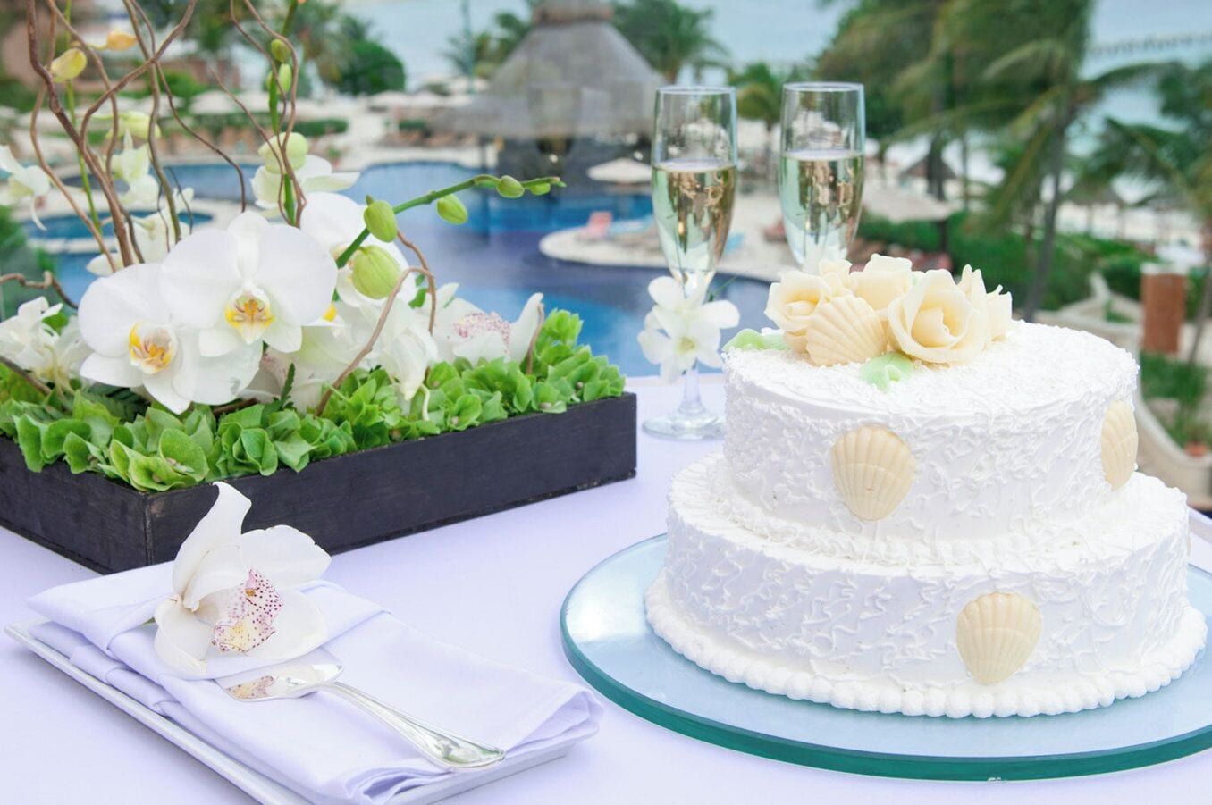 A two-tiered white cake with seashell decorations, placed on a glass cake stand.