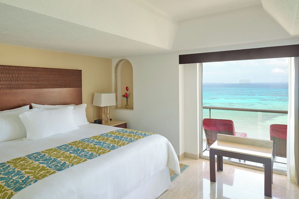 A hotel room with a bed, a nightstand, a lamp, a vase with a flower, and a balcony with a view of the ocean.
