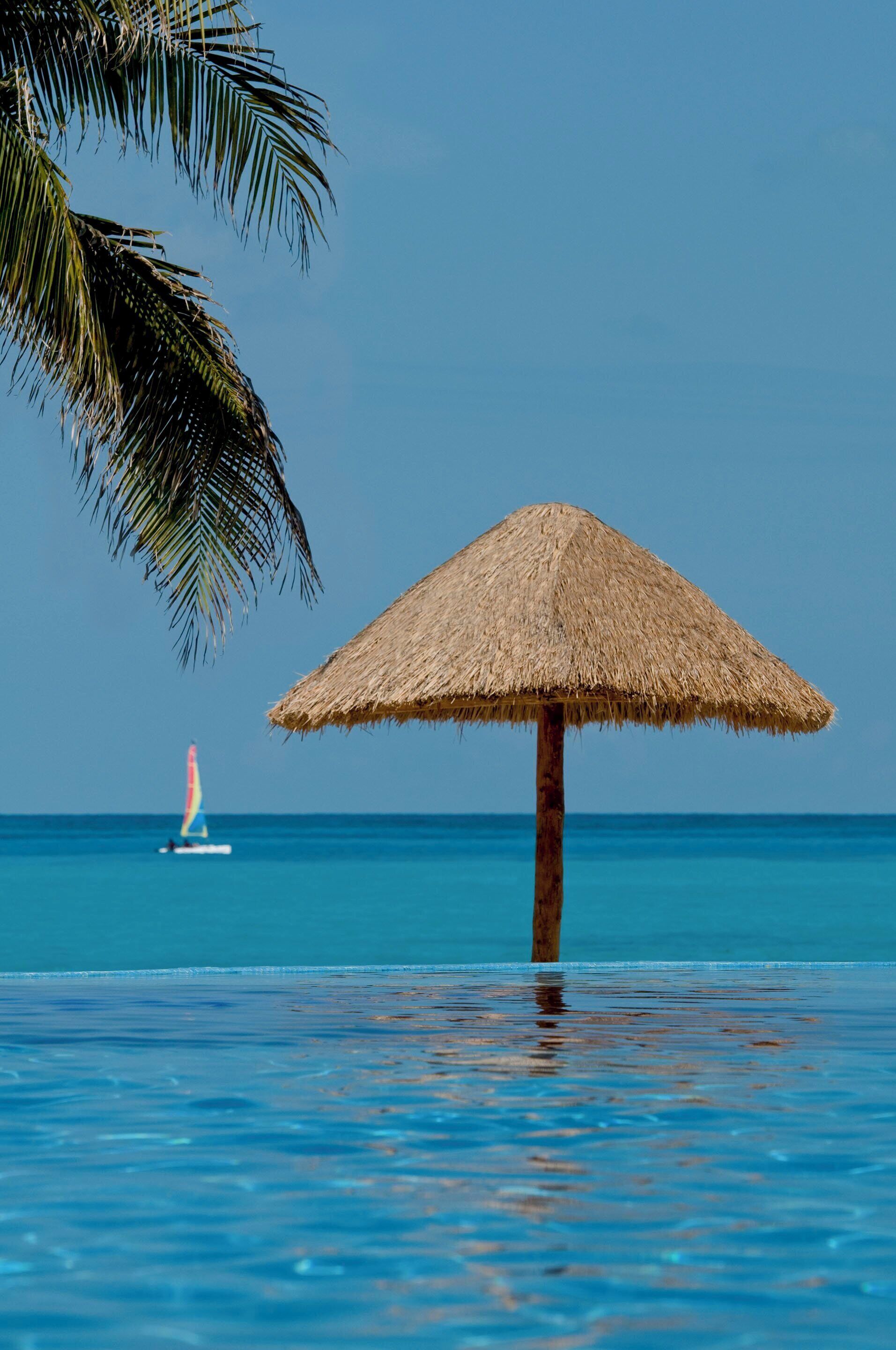 A thatched umbrella in a pool with a clear view of the ocean and a sailboat in the distance.