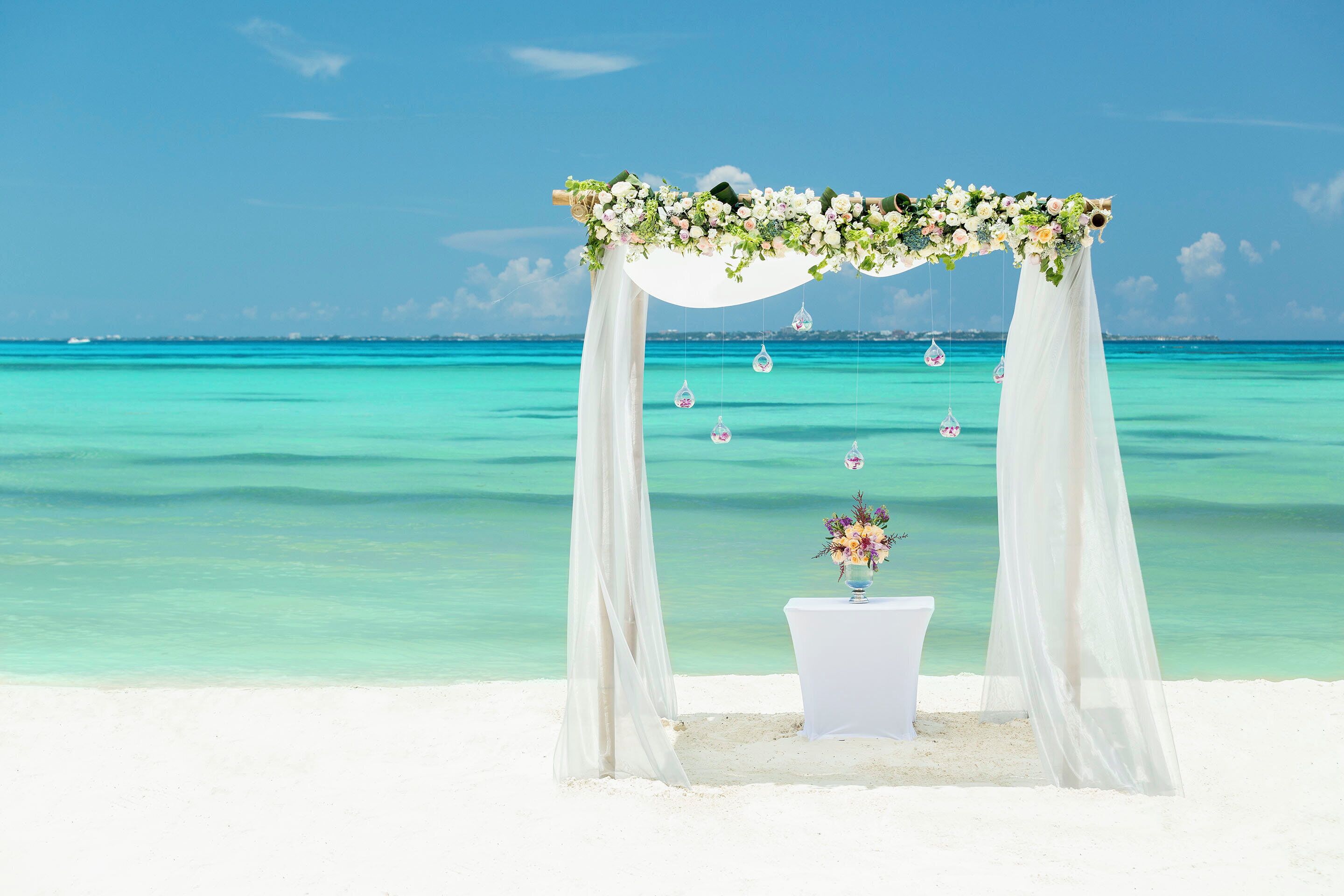 A beach wedding arch with white drapes and floral decorations, a white table with a floral arrangement, and a clear turquoise sea in the background.