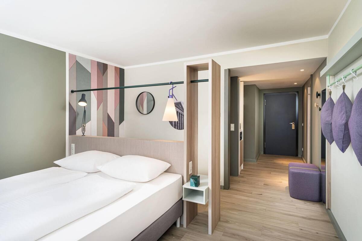 A modern hotel room with a bed, bedside table, and a mirror. There is a hallway leading to another room.