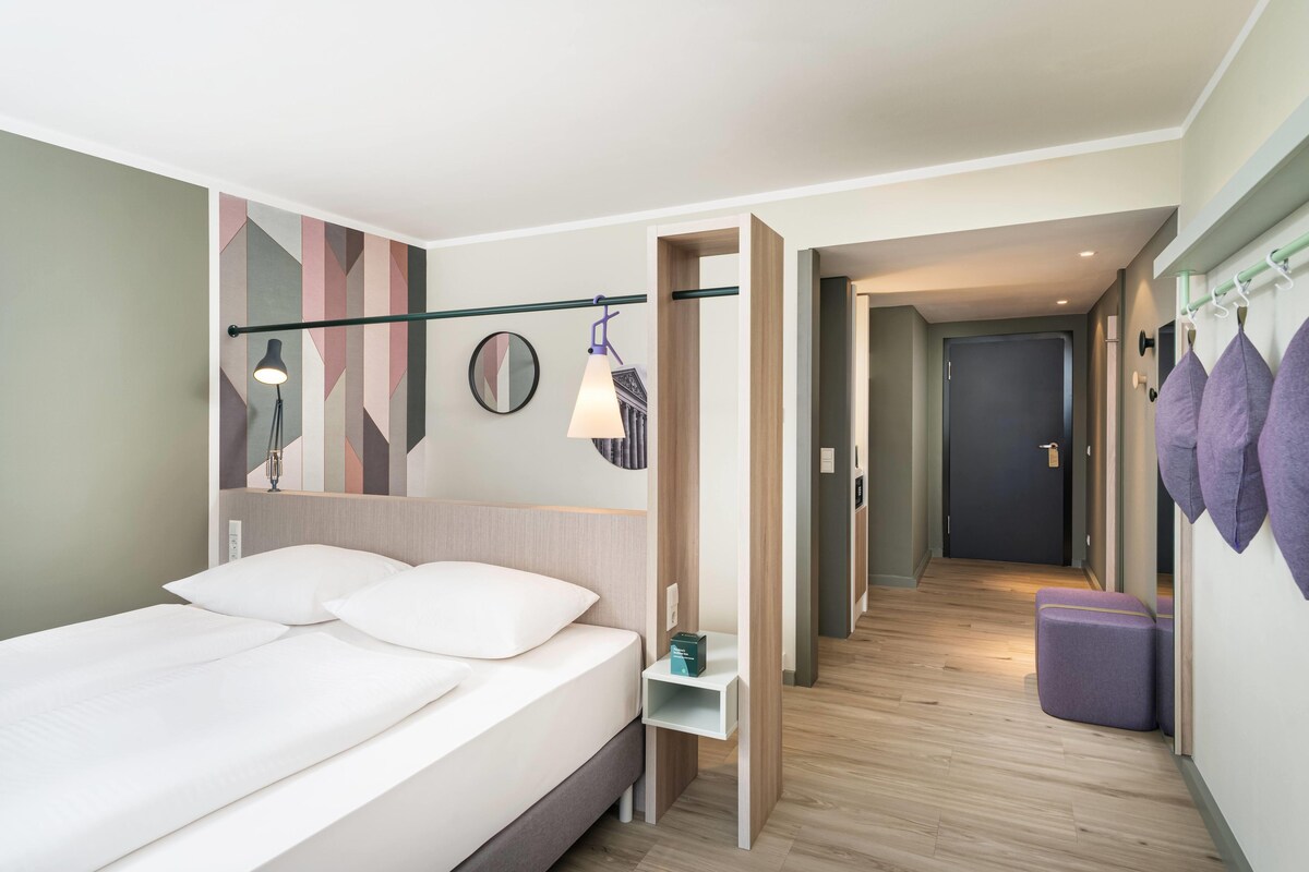 Ein modernes Hotelzimmer mit Bett, Nachttisch, Lampe und Blick in die Hotellobby.