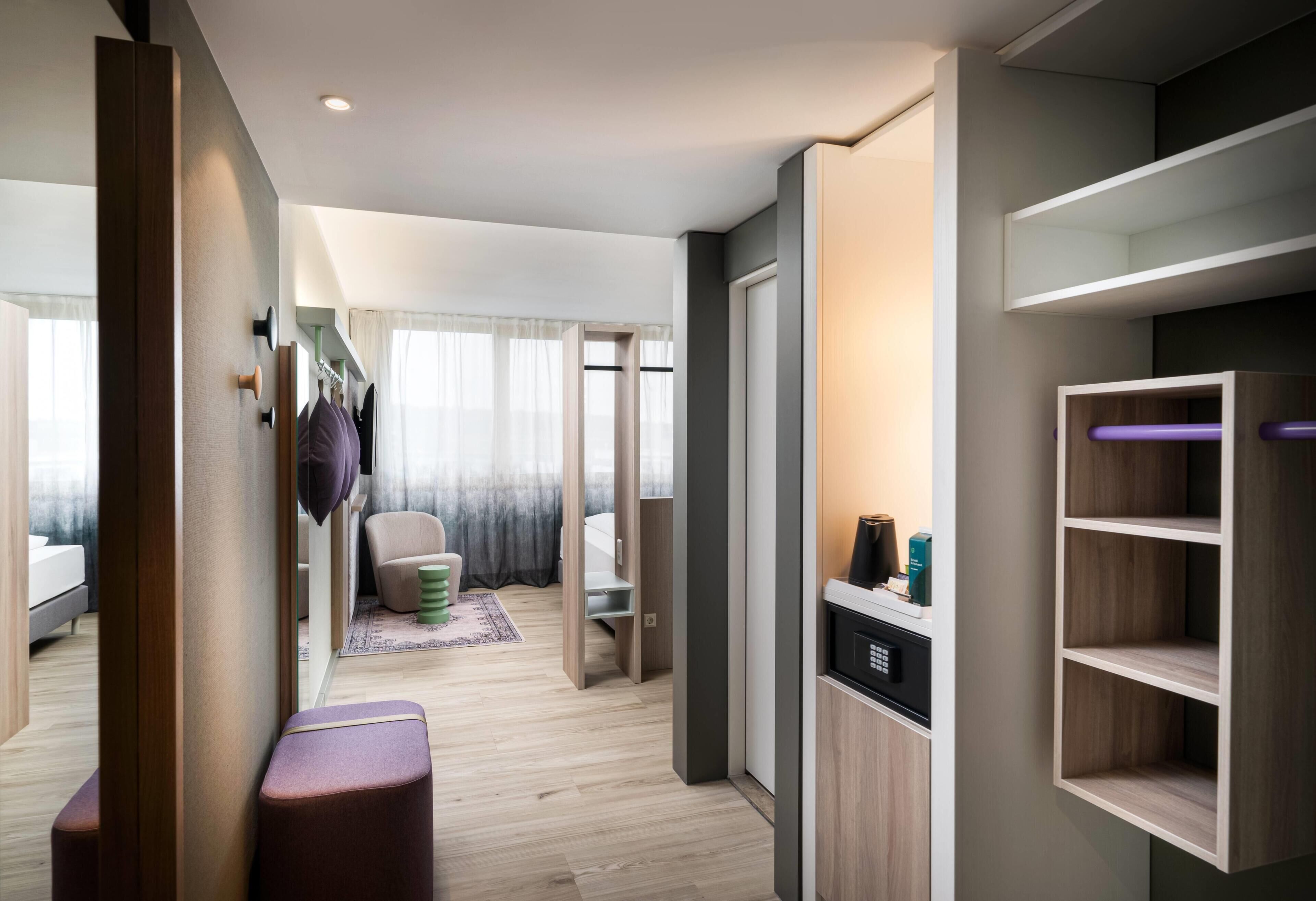 Ein modernes Hotelzimmer mit Holzboden, einer Sitzecke mit Sessel und Hocker, einem eingebauten Kleiderschrank und einer kleinen Küchenzeile.