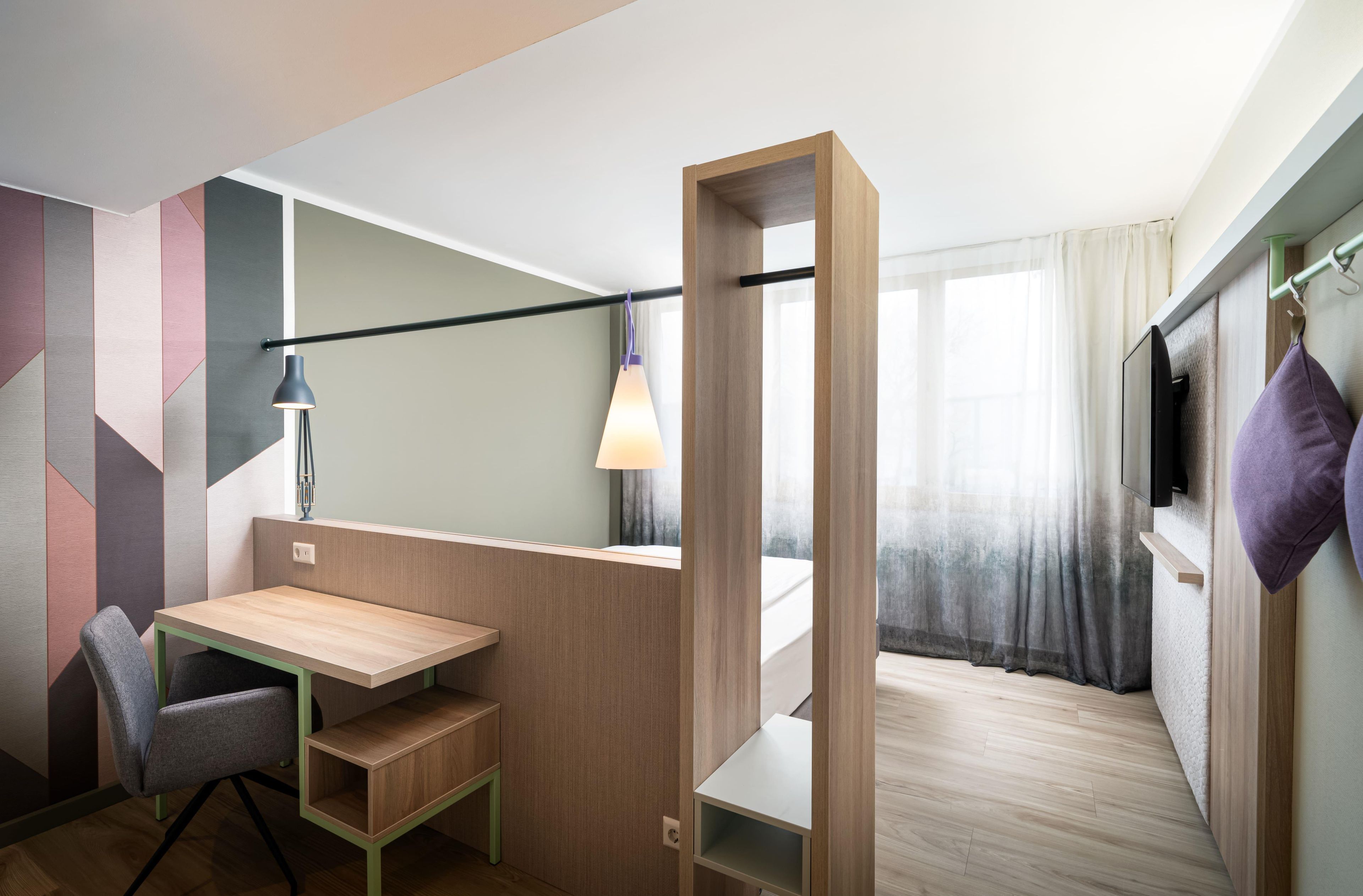 Une chambre d’hôtel de taille réduite, équipée d’un bureau, d’une chaise et d’un lit avec une tête de lit.