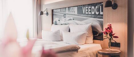 A modern bedroom with a large bed, a headboard with a racing car poster, a bedside table with a vase, and a wall-mounted lamp.