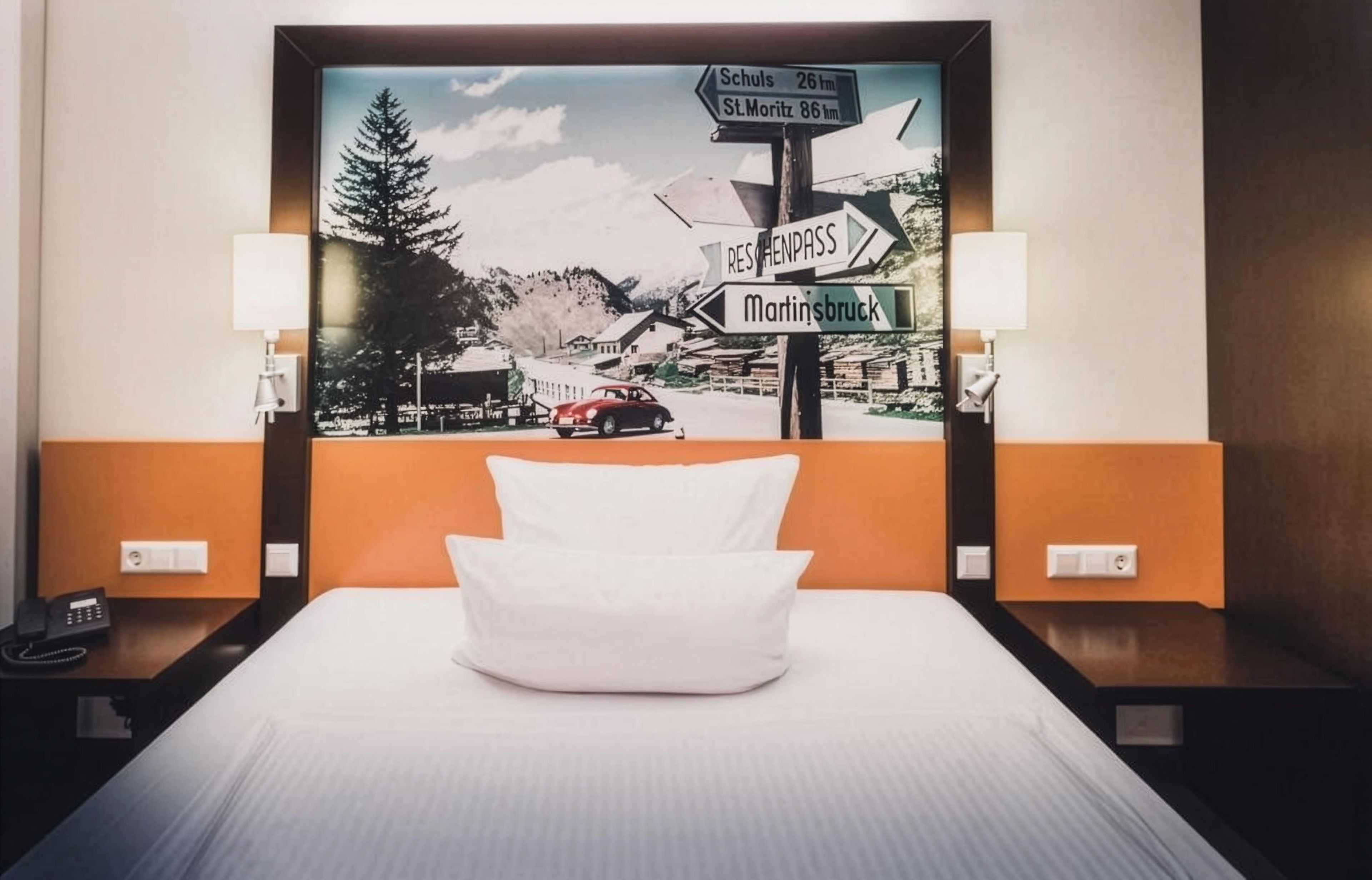 A hotel room with a bed, a nightstand with a phone, and a wall with a framed picture of a street sign.
