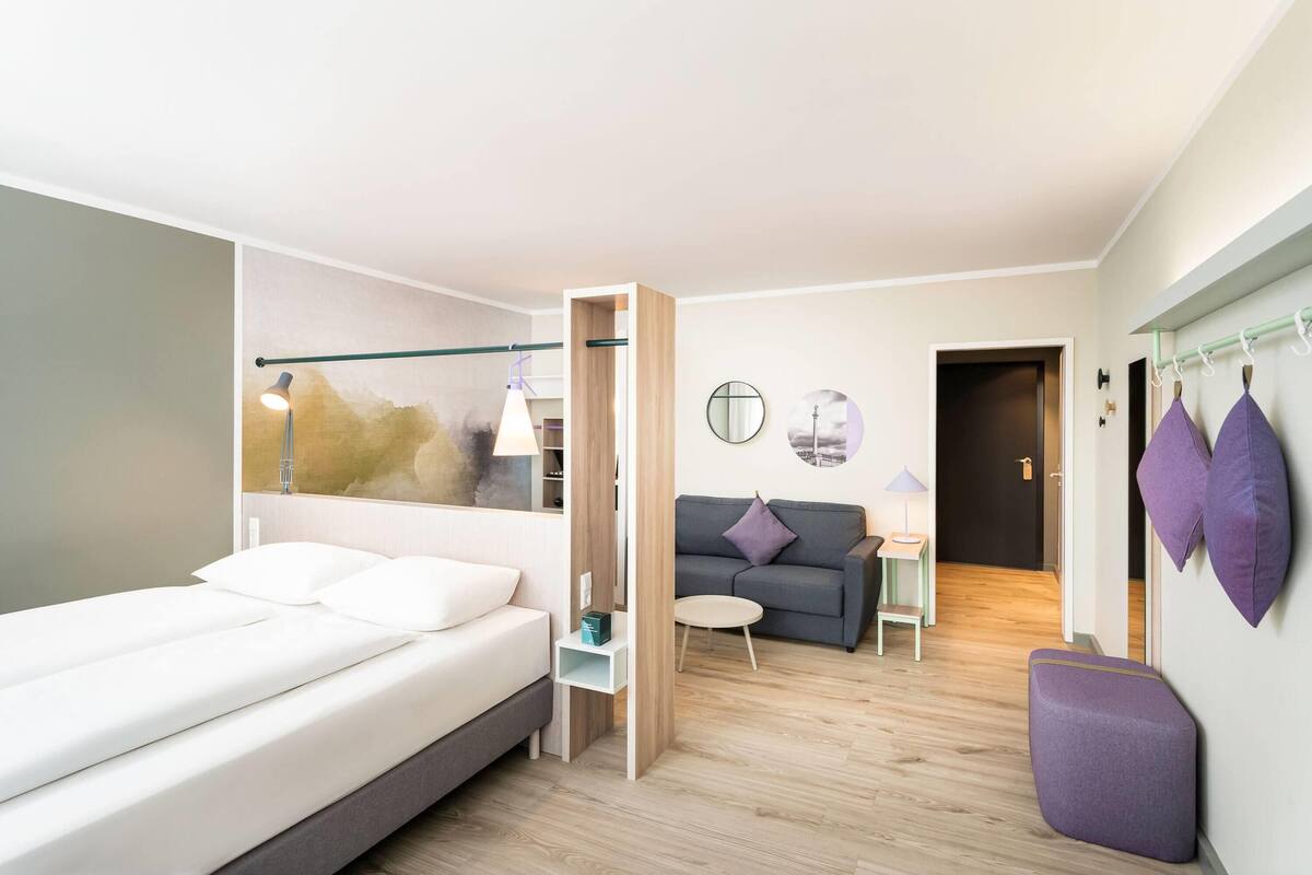 A modern hotel room with a bed, a small sofa, a side table, and a wall-mounted shelf.