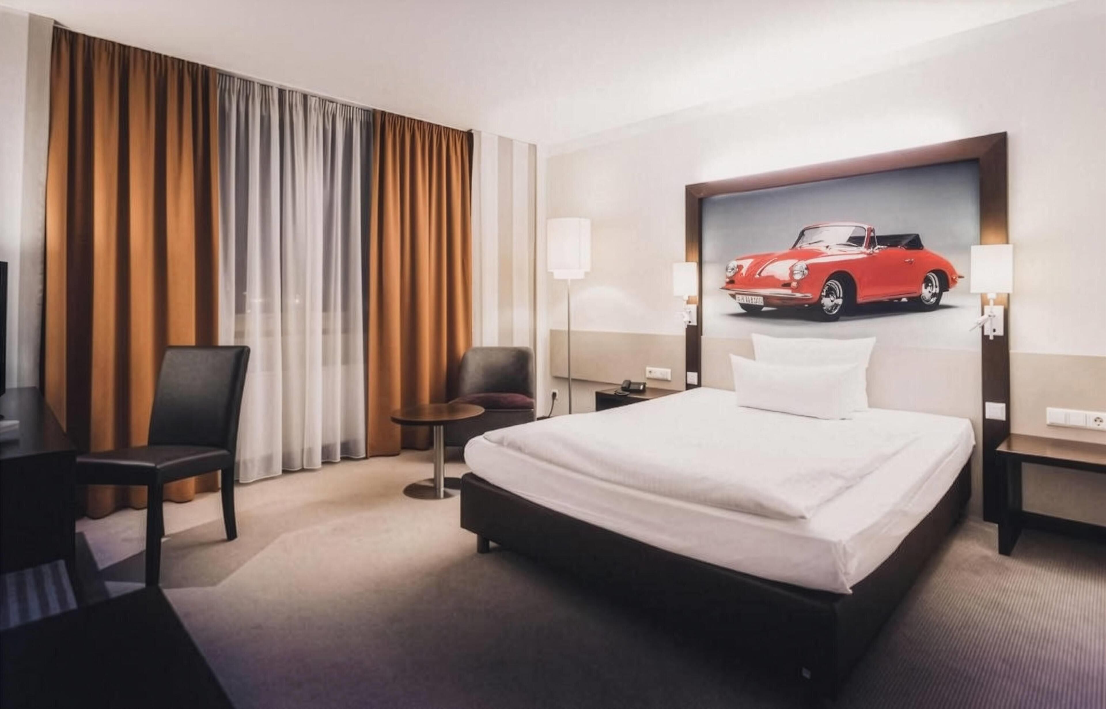 A hotel room with a bed, a desk, a chair, a nightstand, and a wall-mounted lamp.