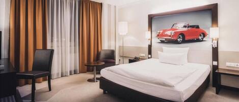 A hotel room with a bed, a desk, a chair, a nightstand, and a wall-mounted lamp.