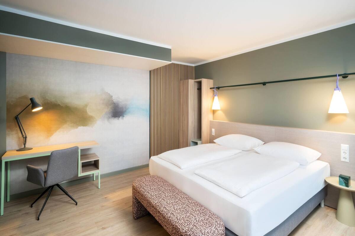A modern hotel room with a bed, desk, chair, bench, and artwork on the wall.