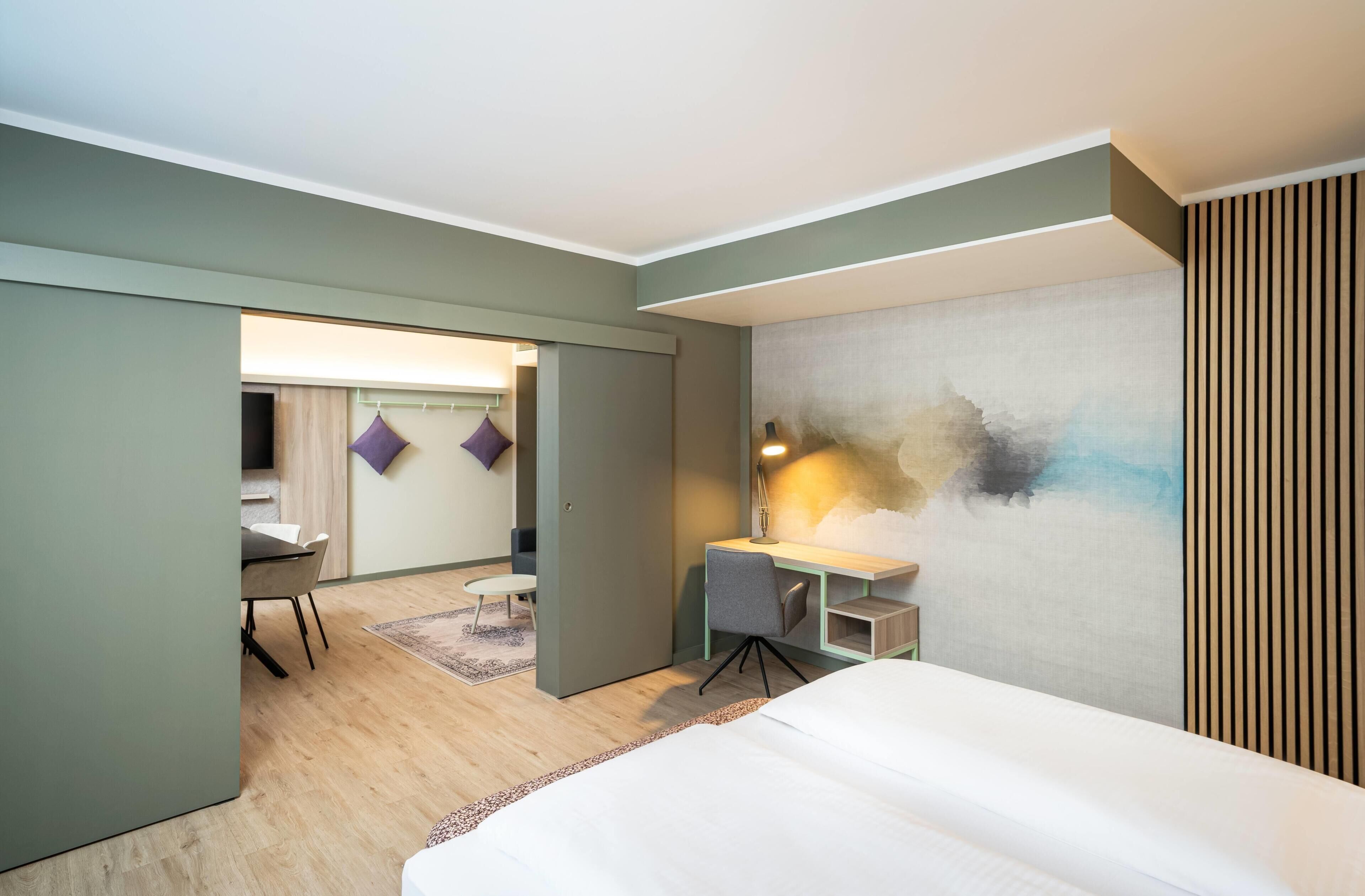 A modern hotel room with a bed, a desk, and a chair. There is a view of the hallway and another room.