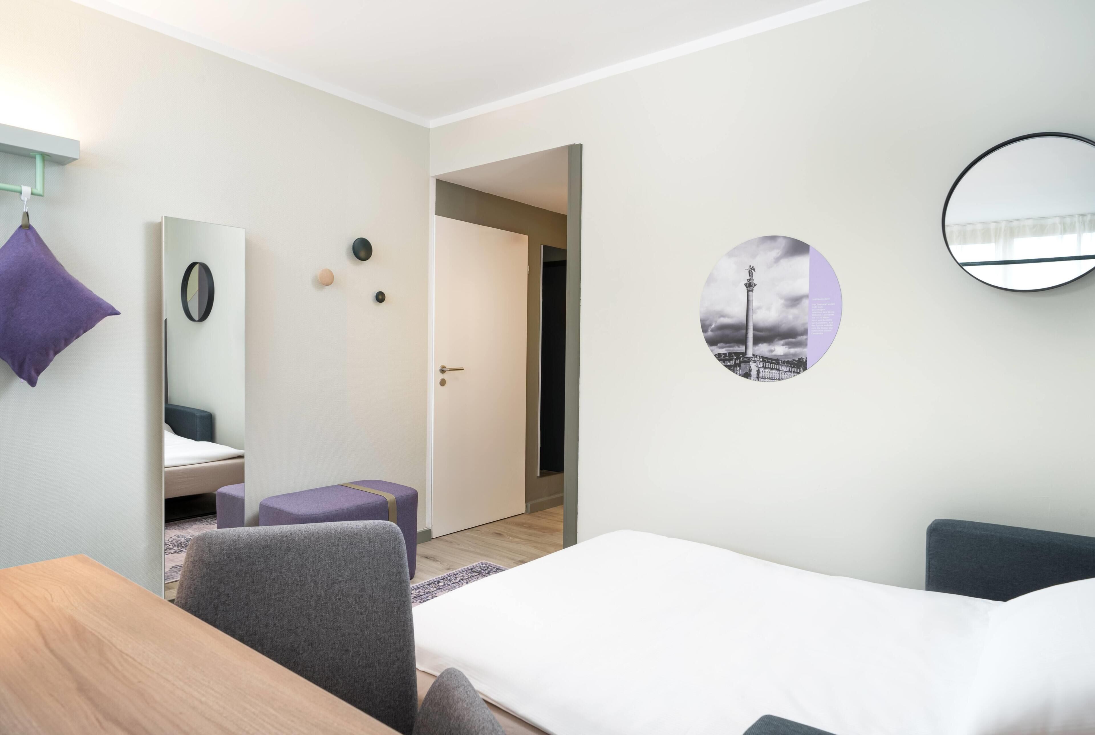A hotel room with a bed, a desk, a chair, a mirror, and a wall with a circular picture.