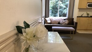 Una mesa de mármol con un jarrón de cristal transparente que contiene flores blancas, un sofá marrón oscuro con un cojín de color claro y una cocina con armarios de madera y un microondas.