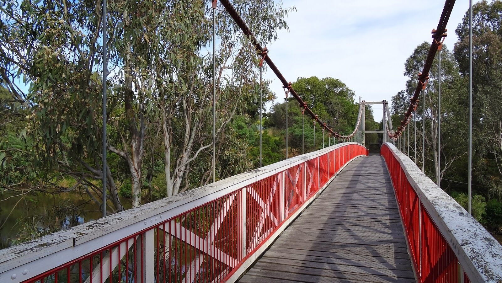 Un puente colgante con barandilla roja y pasarela de madera, rodeado de árboles y una masa de agua.