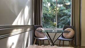Habitación de esquina con mesa de vidrio, dos sillas, un banco mullido, un cuadro enmarcado y una ventana con cortinas.
