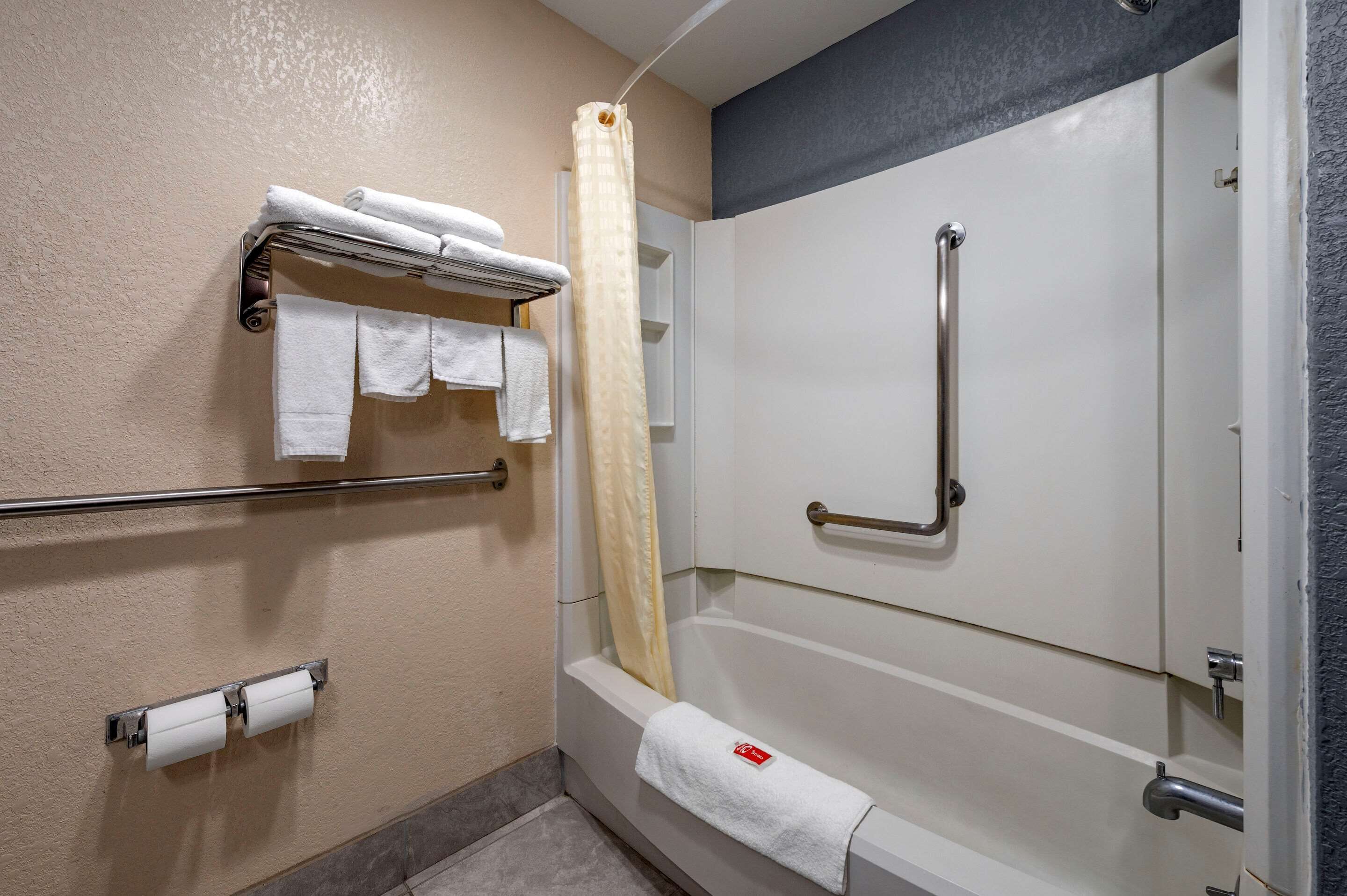 A bathroom with a bathtub, a shower, and a handrail. There are towels and toilet paper on the wall.