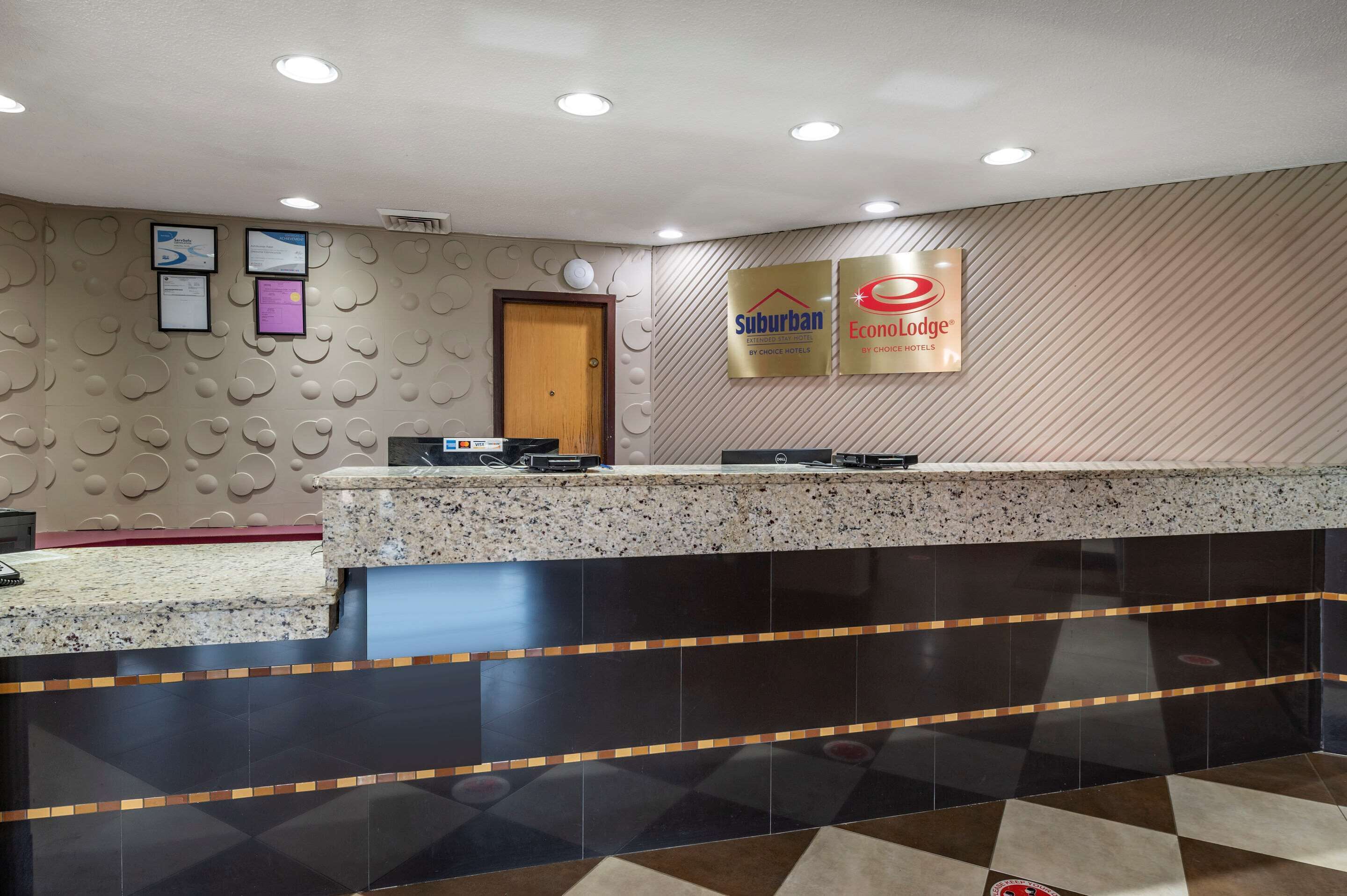 Zona de recepción de un hotel con un mostrador de mármol, dos certificados enmarcados en la pared, y carteles para los hoteles 'Suburban' y 'Economy Lodge'.