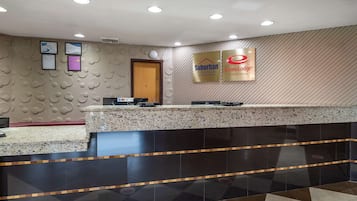 Zona de recepción de un hotel con un mostrador de mármol, dos certificados enmarcados en la pared, y carteles para los hoteles 'Suburban' y 'Economy Lodge'.