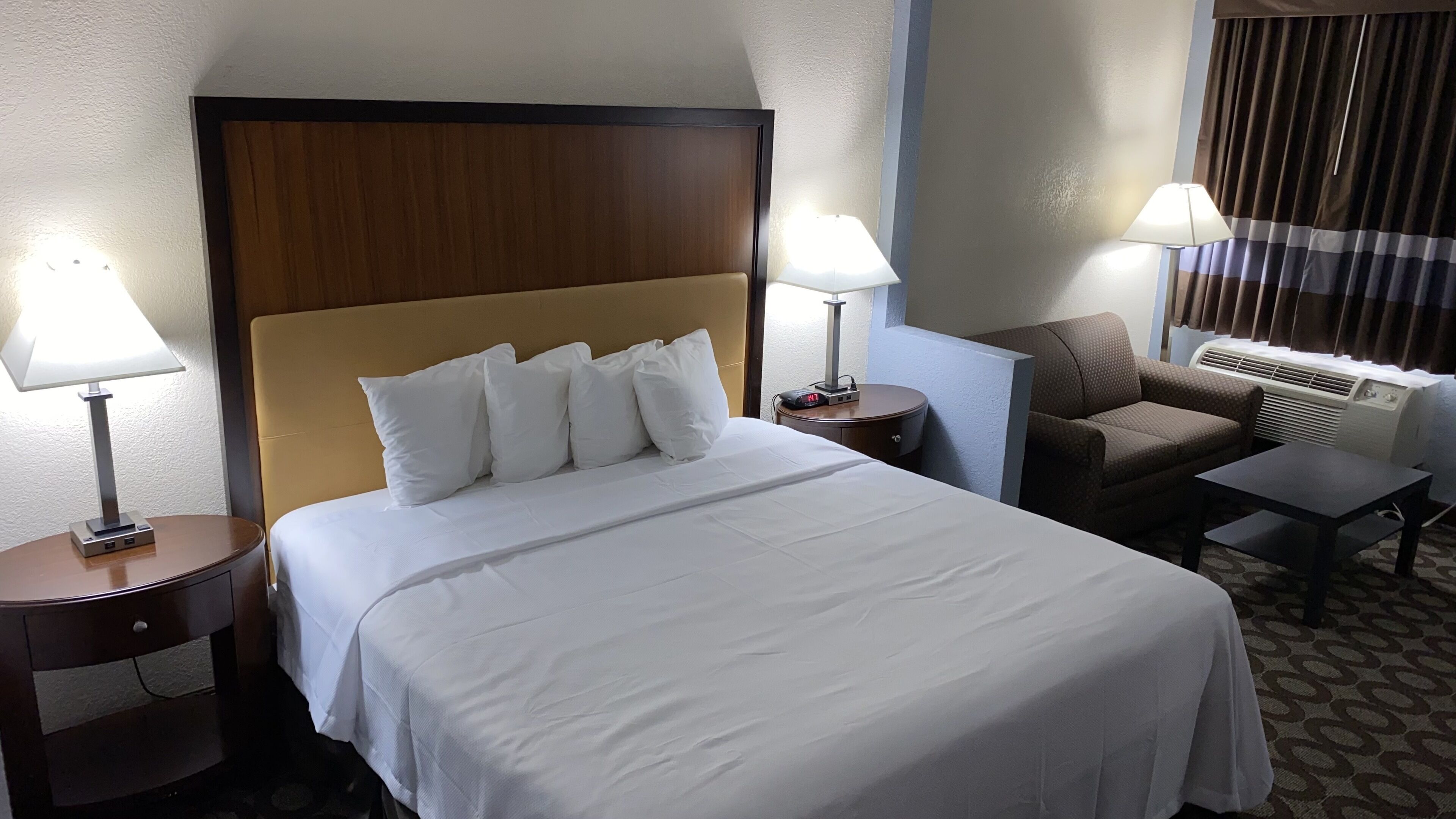 A hotel room with a large bed, two bedside tables with lamps, a chair, a small table, and an air conditioning unit.