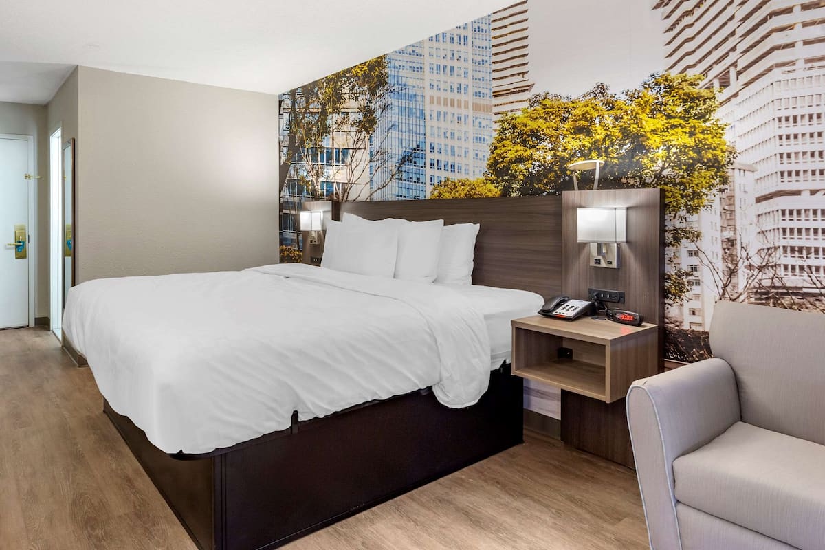 A hotel room with a large bed, a nightstand with a phone and lamp, a chair, and a cityscape mural on the wall.