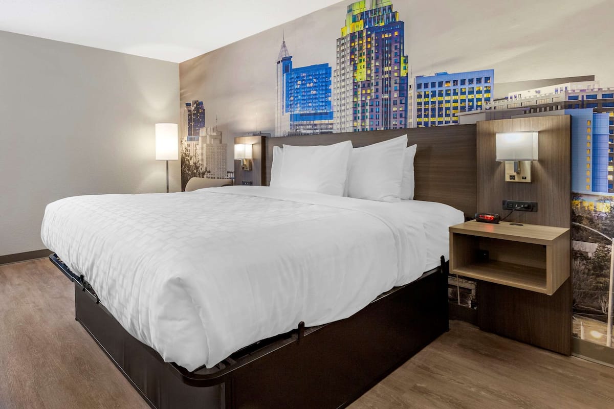 A hotel room with a large bed, a nightstand with a lamp, and a cityscape mural on the wall.