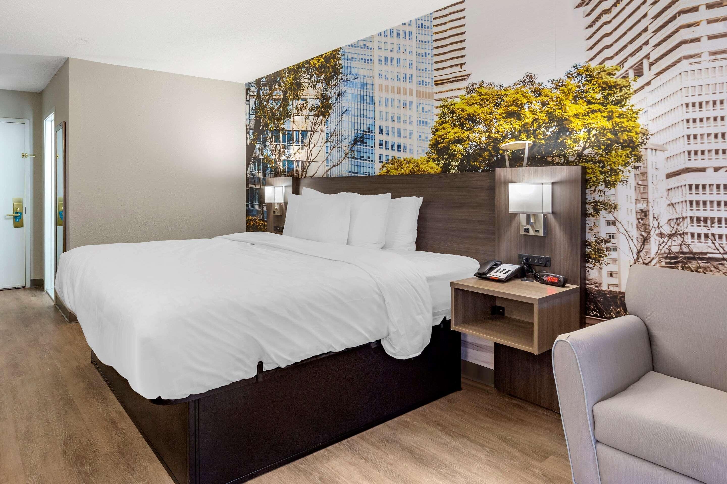 A hotel room with a large bed, a nightstand with a phone and lamp, a chair, and a cityscape mural on the wall.
