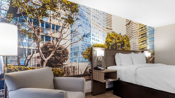 A modern hotel room with a large bed, a seating area, a nightstand, and a wall mural of a cityscape. - Clarion Pointe Raleigh Midtown (Raleigh)