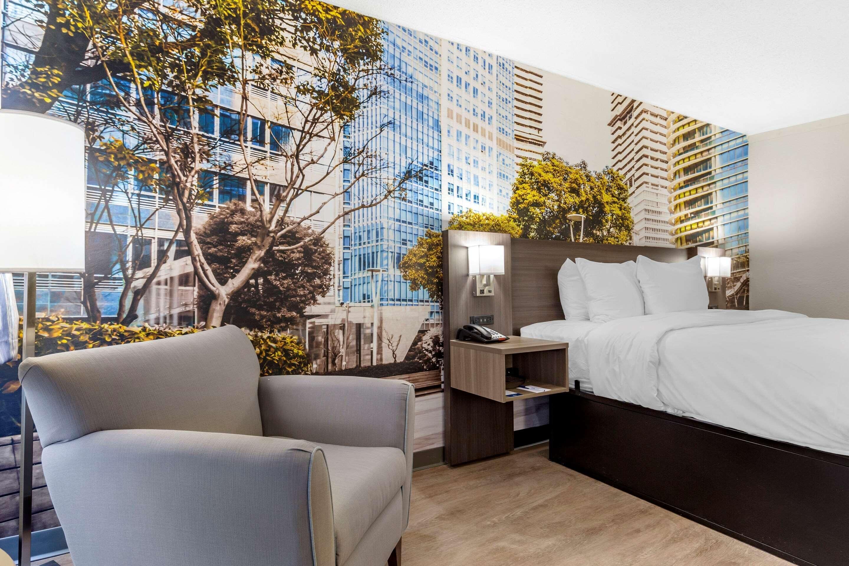A modern hotel room with a large bed, a chair, a nightstand with a lamp, and a wall mural of a cityscape.