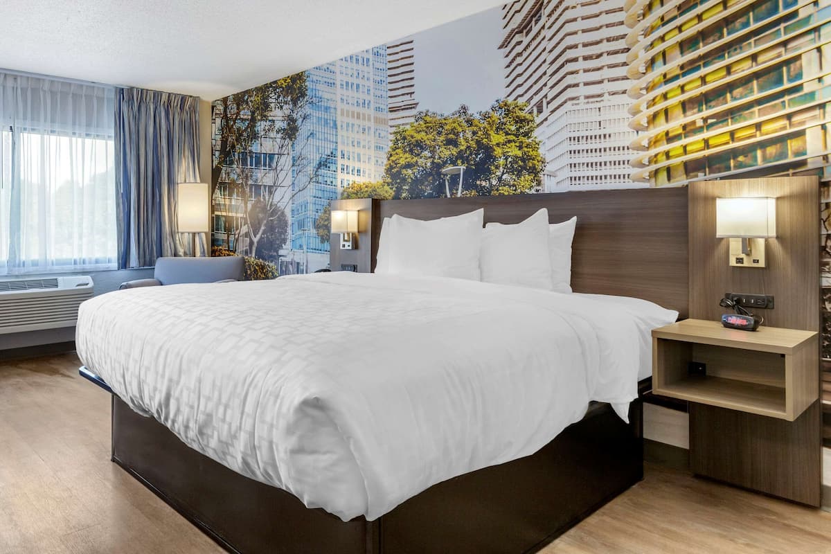 A hotel room with a large bed, a nightstand, a wall-mounted lamp, and a window with curtains.