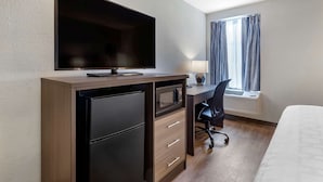 A hotel room with a flat-screen TV mounted on the wall, a desk with a chair, a refrigerator, a microwave, a window with curtains, and a bed. - Clarion Pointe Raleigh Midtown (Raleigh)